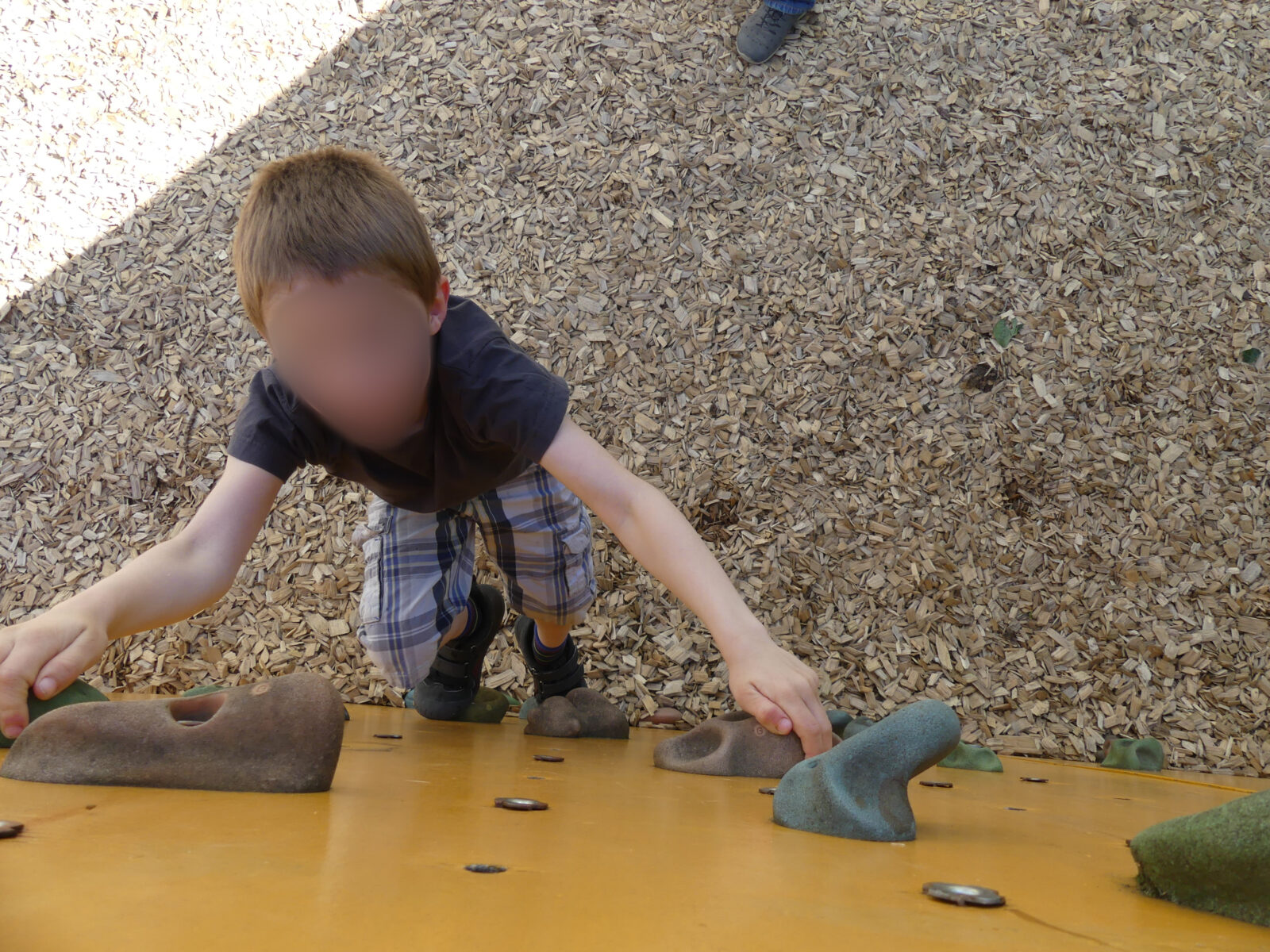 Ein kleines Kind klettert an einer Kletterwand mit bunten Griffen nach oben. Das Kind trägt ein dunkelgraues T-Shirt und karierte Shorts. Der Boden ist mit Holzspänen bedeckt, die eine weiche Landung gewährleisten.