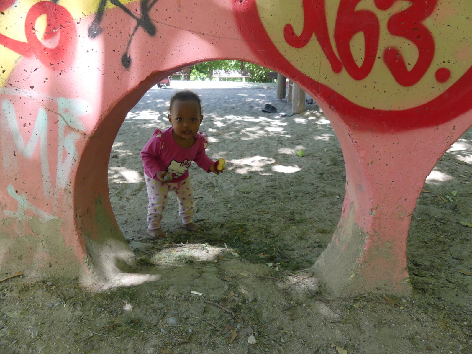 Ein kleines Kind spielt in einem Park und schaut durch eine große, bunt bemalte Betonröhre. Die Röhre ist mit Graffiti verziert und die Umgebung ist schattig und bewaldet. Das Kind trägt ein pinkes Oberteil mit einer Hello Kitty Figur darauf und hält etwas in den Händen.