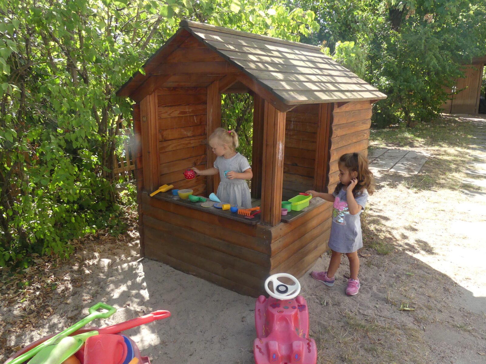 Zwei kleine Kinder spielen in einem hölzernen Spielhaus im Freien. Das Spielhaus ist mit verschiedenen bunten Spielzeugen ausgestattet, darunter Geschirr und Küchenutensilien. Im Vordergrund ist ein rotes Spielzeugauto auf sandigem Boden zu sehen, während die Umgebung von grünen Bäumen und Sträuchern umgeben ist.