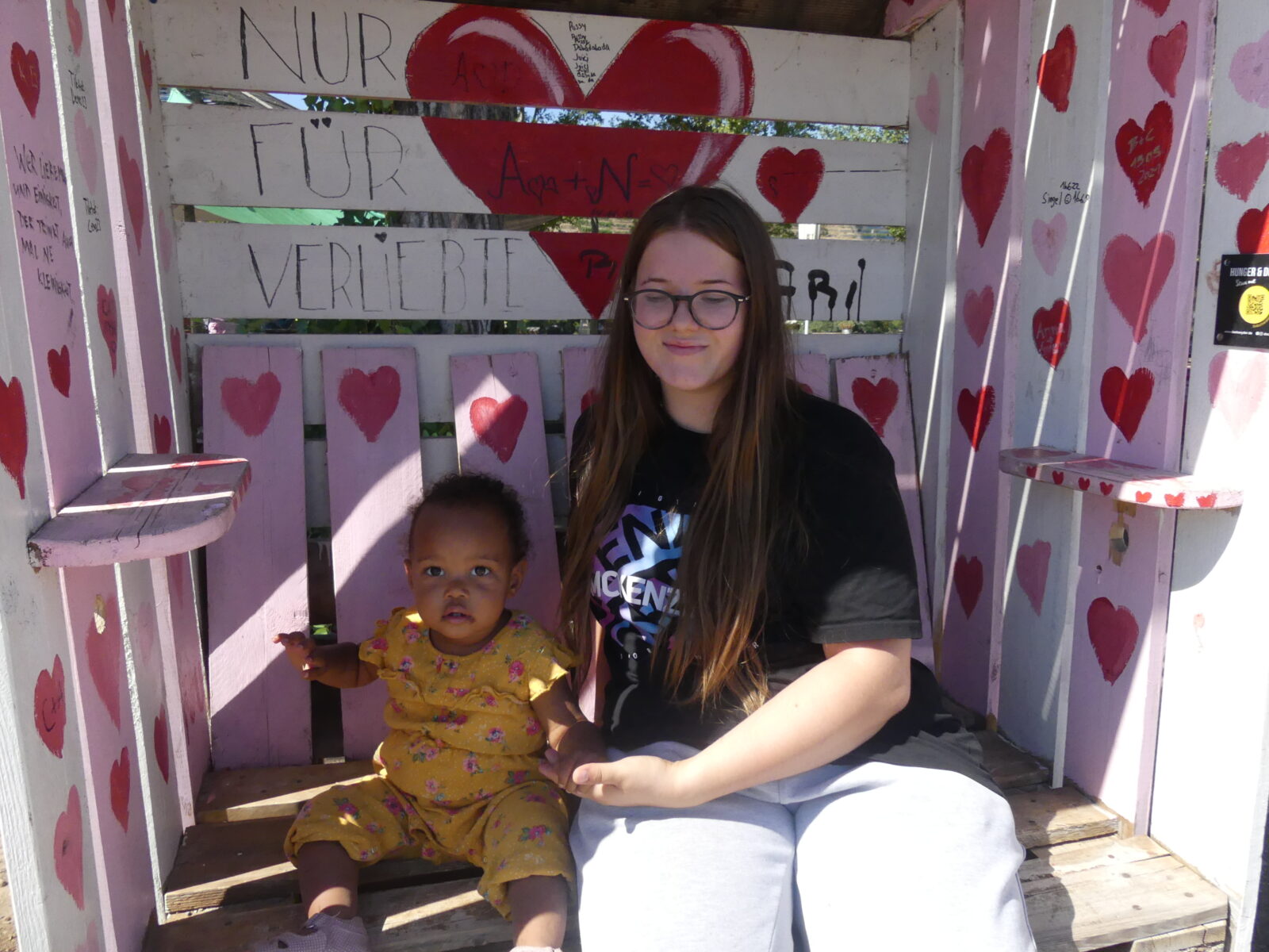 Eine Frau sitzt mit einem kleinen Kind auf einer bunt bemalten Bank mit Herzen und der Aufschrift "Nur für Verliebte".