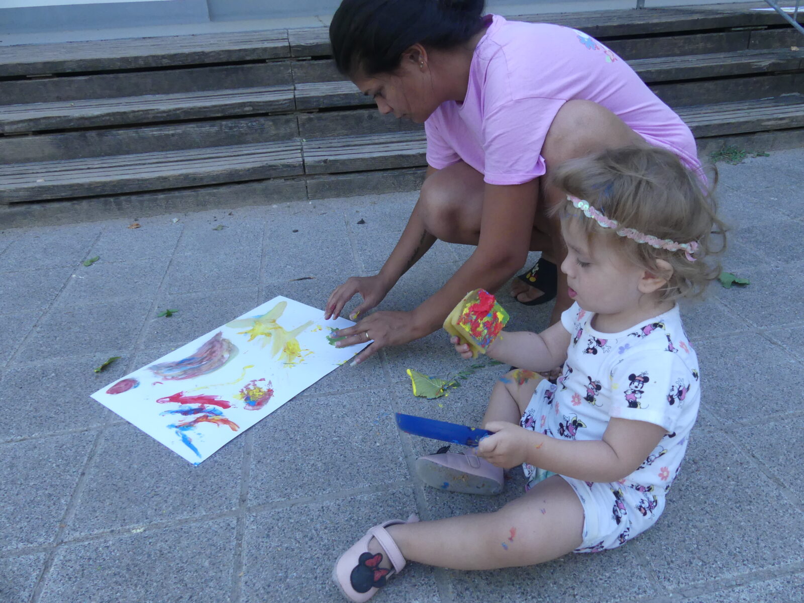 Ein Kind und eine erwachsene Person malen gemeinsam mit Fingerfarben auf einem Blatt Papier, während sie draußen auf dem Boden sitzen. Das Kind hält einen Farbbehälter und einen Pinsel in den Händen.