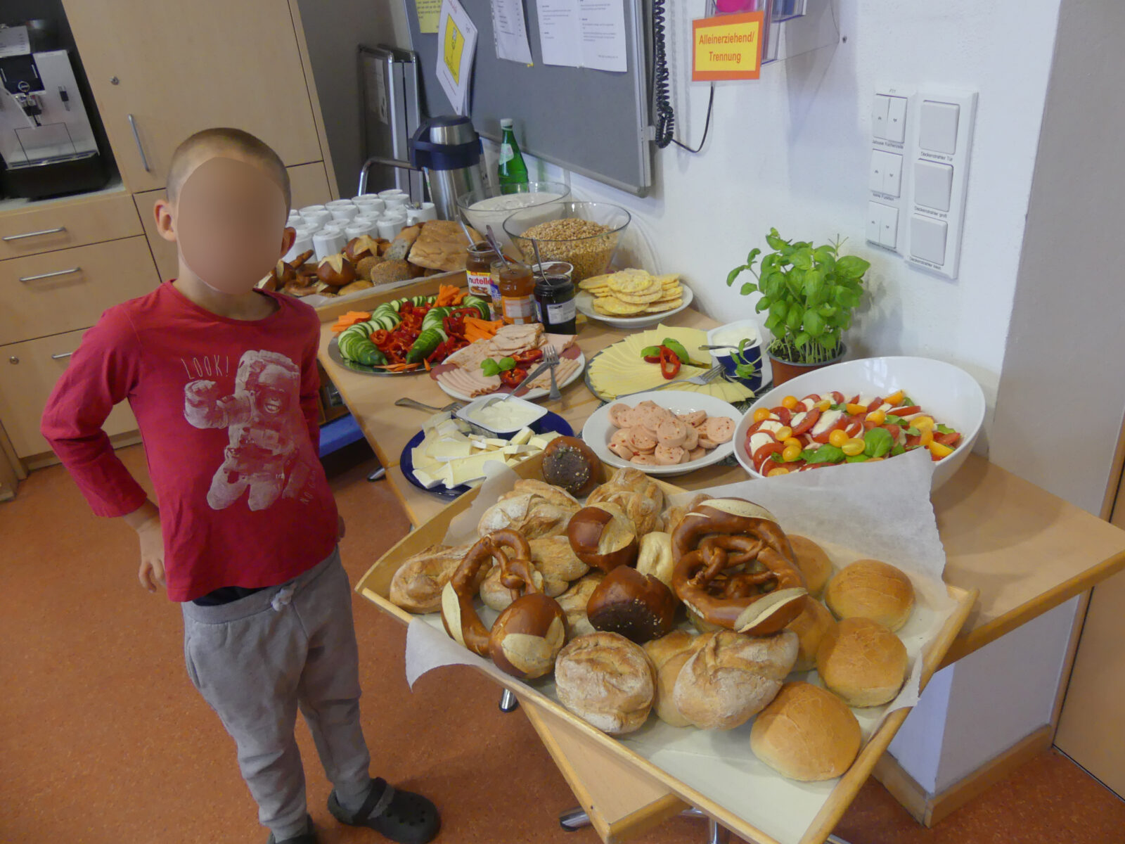 Ein Kind steht vor einem Tisch mit einer Auswahl an frischen Brötchen, Brezeln, Käse, Wurst, Gemüse und Salat in einer Küche oder einem Gemeinschaftsraum. Im Hintergrund sind weitere Lebensmittel, Getränke und Küchenutensilien zu sehen.