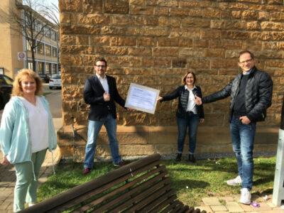 Eine Gruppe von vier Personen steht vor einer Steinmauer im Freien. Zwei der Personen halten gemeinsam ein großes Zertifikat, während die anderen daneben stehen. Im Hintergrund sind ein Gebäude und parkende Autos zu sehen.