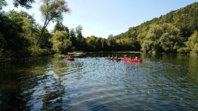 Eine Gruppe von Menschen paddelt in roten Kanus auf einem ruhigen Fluss, umgeben von üppiger grüner Vegetation und Bäumen. Der Himmel ist klar und blau, und die Szenerie vermittelt ein Gefühl von Frieden und Naturverbundenheit. Im Hintergrund sind bewaldete Hügel zu sehen, die die idyllische Landschaft abrunden.