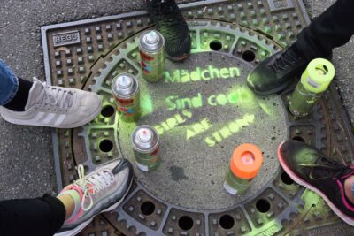 Vier Paar Schuhe umgeben eine bemalte Kanalabdeckung, auf der die Wörter "Mädchen sind cool" und "Girls are strong" in grüner Farbe gesprüht sind. Neben den Schuhen stehen mehrere Spraydosen, die für die Bemalung verwendet wurden. Das Bild vermittelt eine Botschaft der Stärke und Coolness von Mädchen.