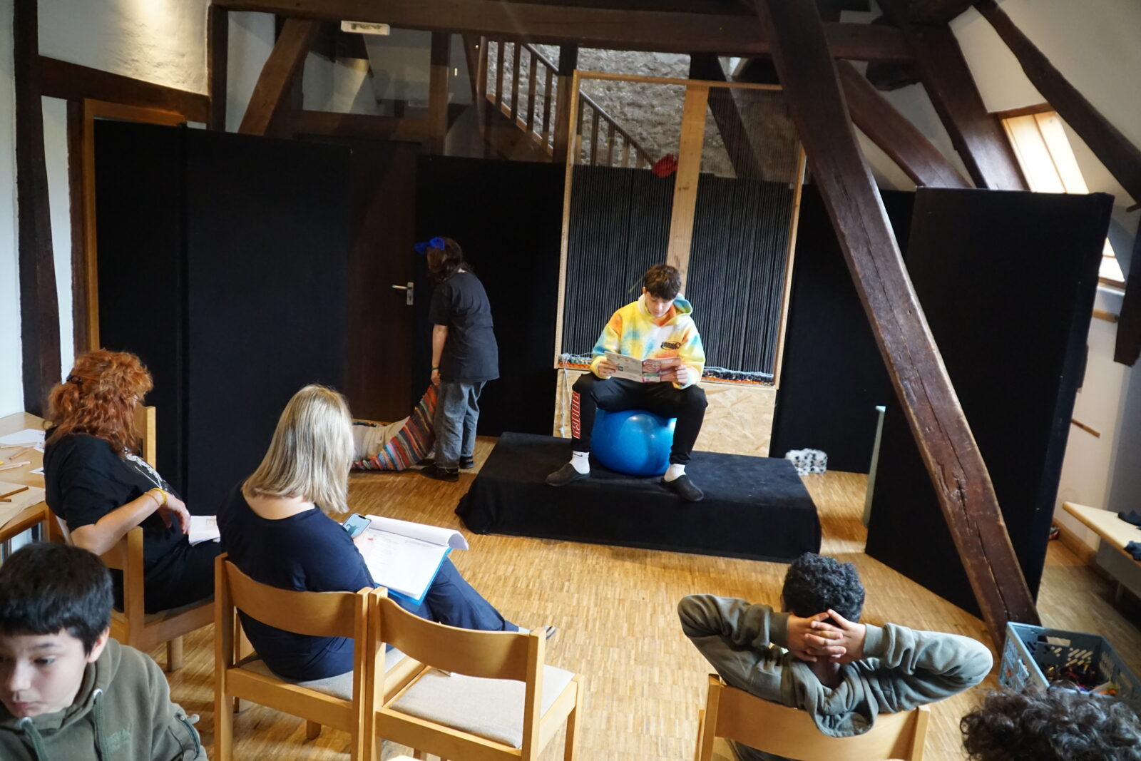 Eine Gruppe von Menschen sitzt in einem Raum, während eine Person auf einem blauen Gymnastikball auf einer kleinen Bühne sitzt und ein Buch liest. Die Umgebung ist rustikal, mit Holzbalken und einem Holzboden. Im Hintergrund ist eine Person zu sehen, die sich mit einem bunten Teppich beschäftigt.