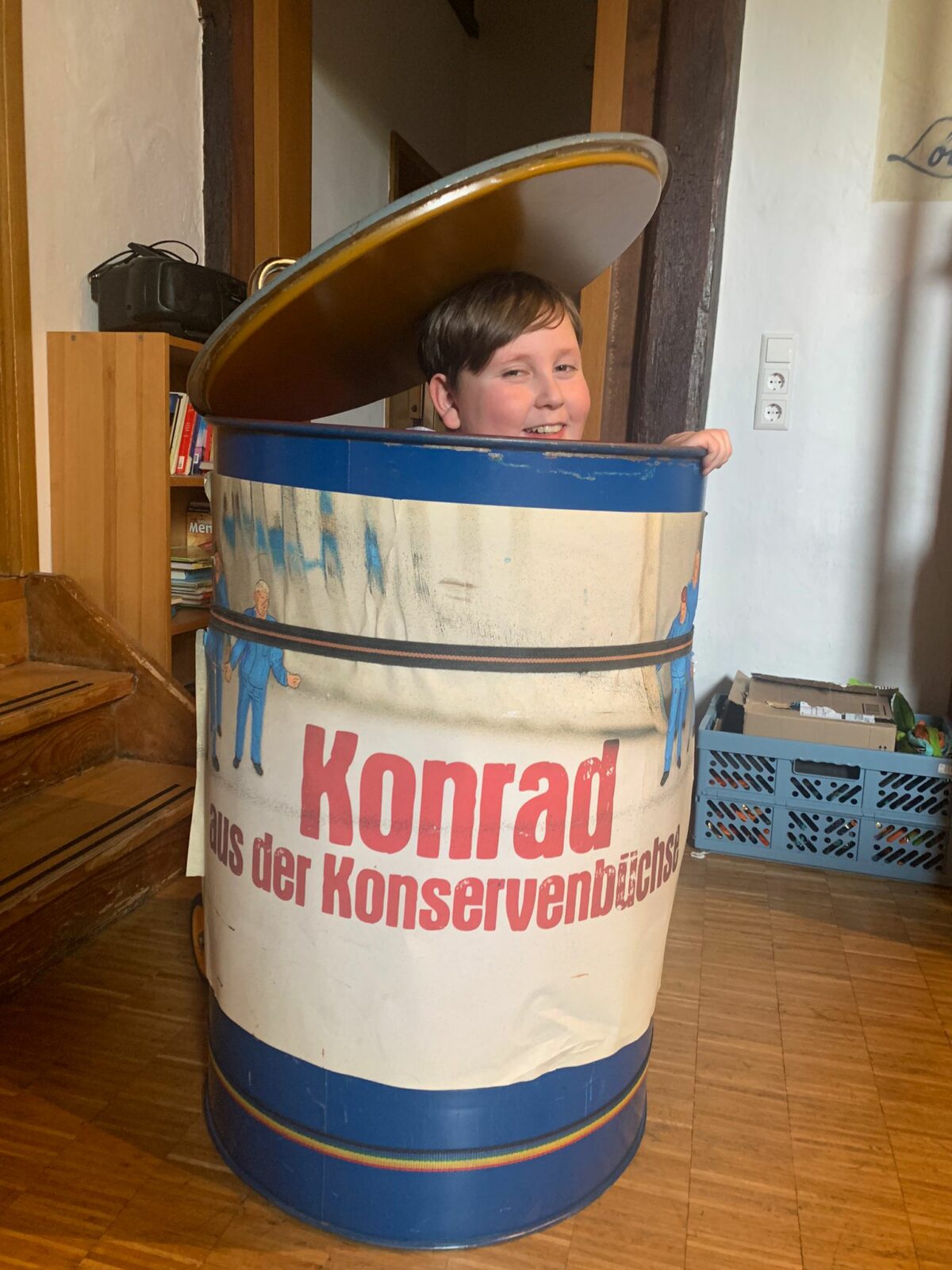 Ein Kind sitzt in einer großen Konservendose, die als Spielzeug verwendet wird. Die Dose hat die Aufschrift "Konrad aus der Konservenbüchse" und ist bunt bemalt. Im Hintergrund sind ein Bücherregal und eine blaue Kiste mit verschiedenen Gegenständen zu sehen.