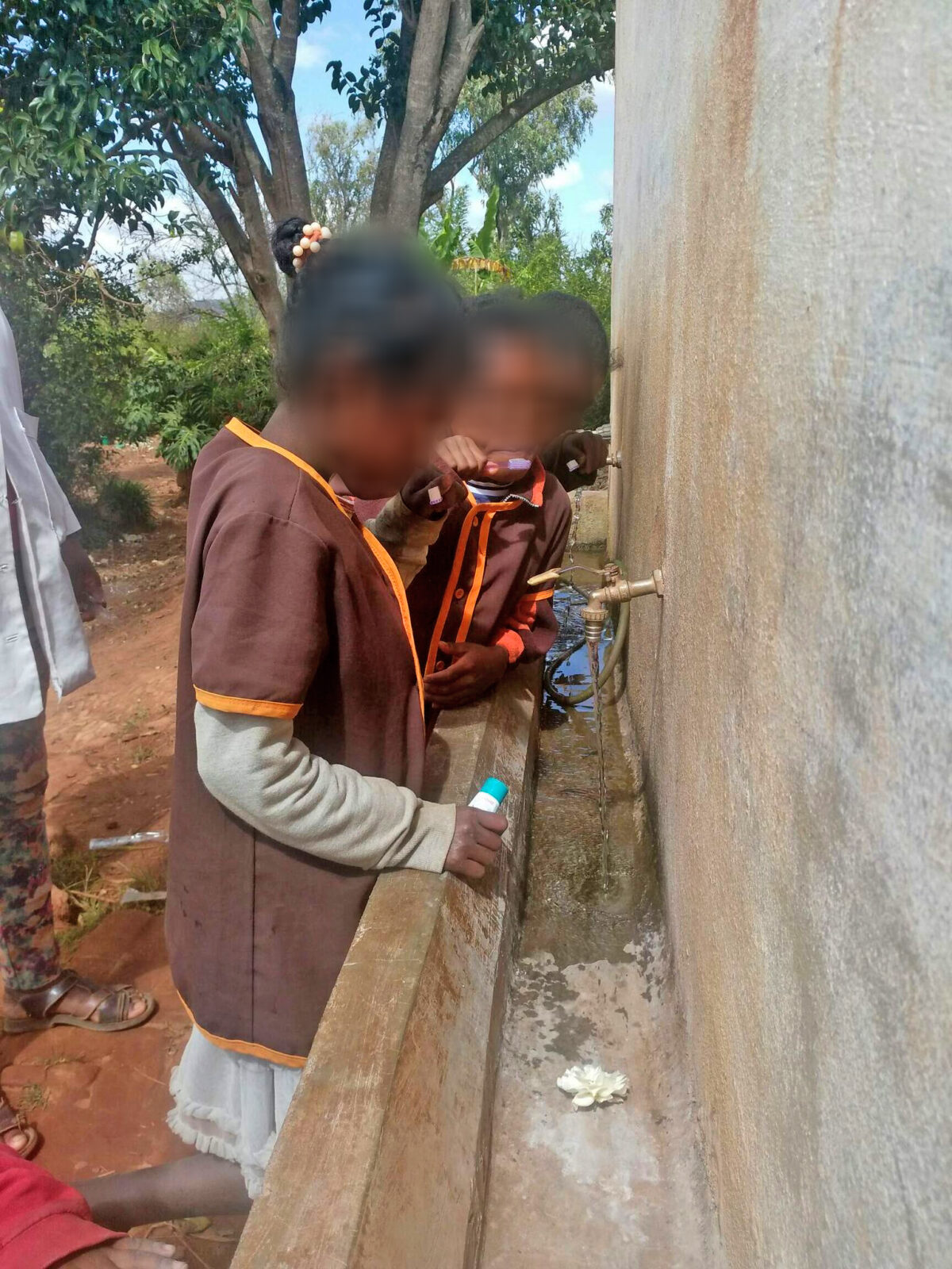 Zwei Kinder stehen an einem Wasserhahn und füllen ihre Flaschen. Sie tragen braune Uniformen mit orangefarbenen Akzenten. Die Umgebung wirkt ländlich und einfach.