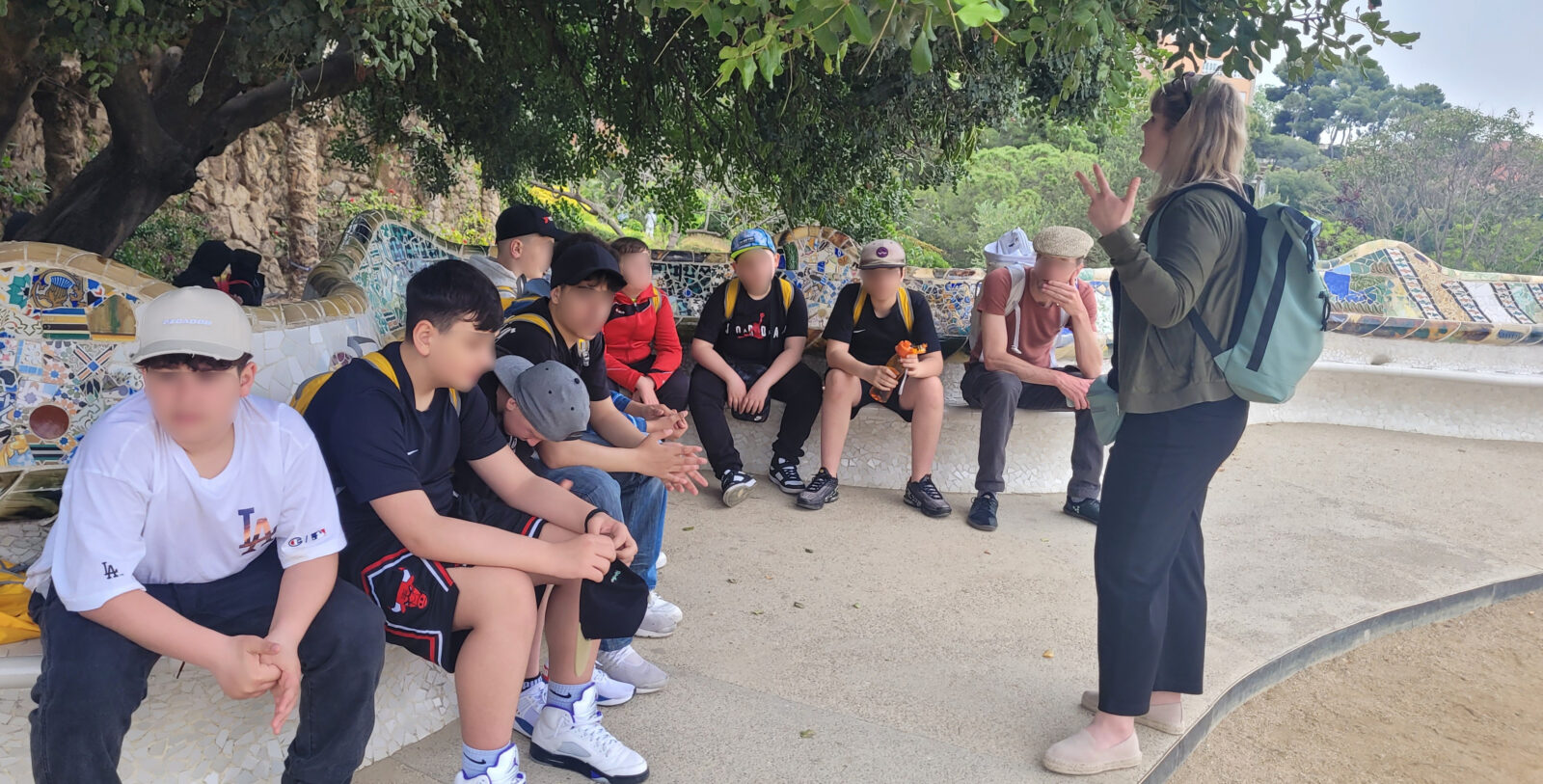 Eine Gruppe von Menschen sitzt auf einer kunstvoll gestalteten Bank im Freien und hört einer Frau zu, die steht und gestikuliert. Im Hintergrund sind Bäume und grüne Landschaft zu sehen.
