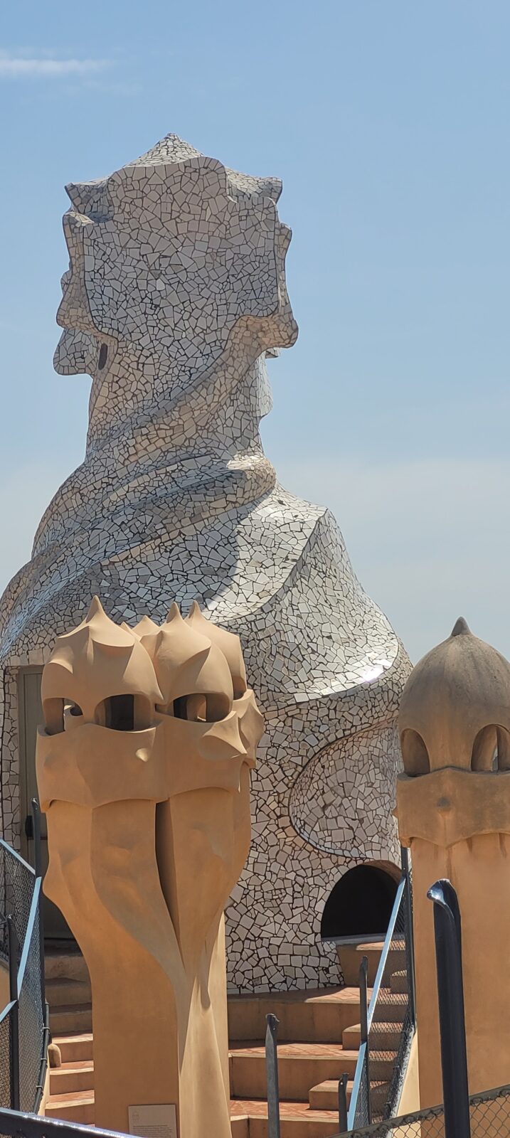 Das Bild zeigt die markanten, skulpturalen Schornsteine und Lüftungstürme auf dem Dach der Casa Milà, einem berühmten Gebäude in Barcelona, entworfen von Antoni Gaudí. Die Türme sind mit Mosaiken aus Keramik und Stein bedeckt und haben organische, wellenförmige Formen, die an natürliche Strukturen erinnern. Im Vordergrund sind zwei sandfarbene Türme zu sehen, während im Hintergrund ein größerer, weißer Turm mit komplexen Mustern hervorsticht. Der Himmel ist klar und blau.