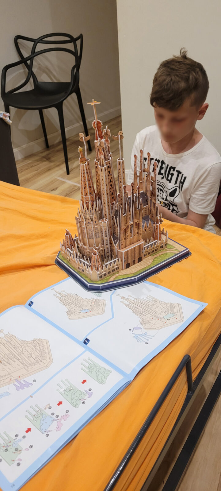 Ein Kind sitzt an einem Tisch und betrachtet ein Modell der Sagrada Familia, das auf einem orangefarbenen Tuch steht. Vor dem Modell liegt eine Anleitung mit Bauplänen. Im Hintergrund steht ein schwarzer Stuhl.