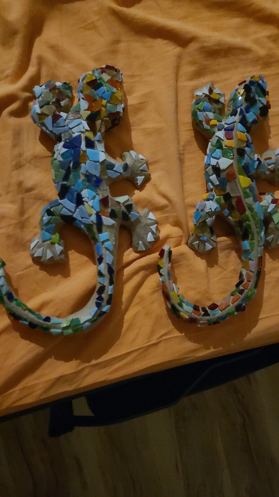 Das Bild zeigt zwei kunstvolle Mosaik-Eidechsen, die auf einem orangefarbenen Tuch liegen. Beide Eidechsen sind mit bunten, geometrischen Steinen verziert, die verschiedene Farben wie Blau, Grün, Gelb und Rot umfassen. Die Eidechsen sind identisch in ihrer Form und Größe, und sie scheinen nebeneinander platziert zu sein.