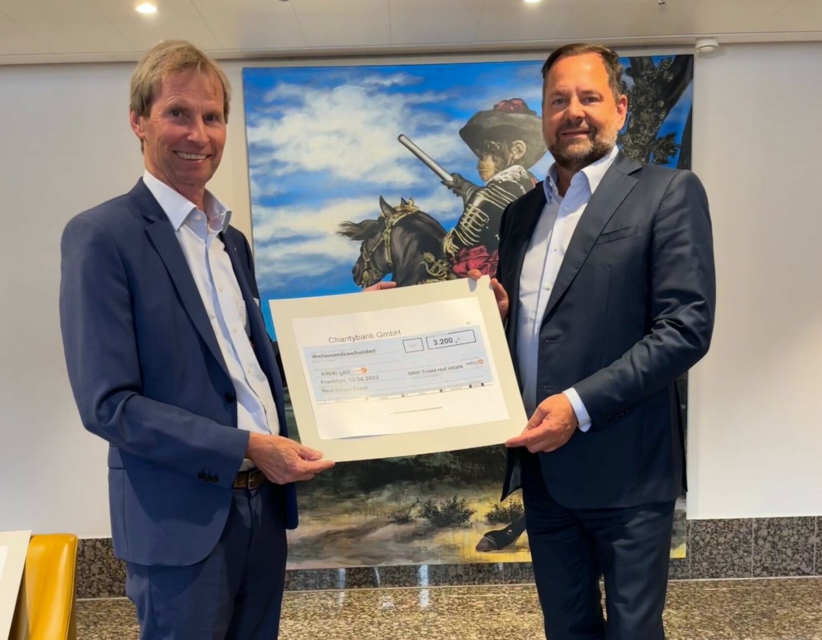 Zwei Männer in Anzügen halten einen großen Scheck über 3.200 Euro vor einem Gemälde, das einen Affen in historischer Kleidung auf einem Pferd zeigt. Der Scheck ist von der Charitybank GmbH und adressiert an die KiNiKi gAG.