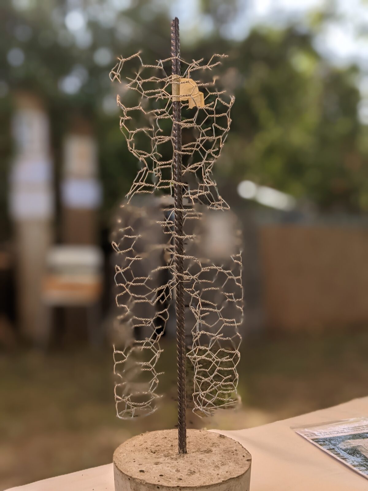 Kunstinstallation aus Drahtgeflecht, das eine menschliche Figur darstellt, befestigt an einer vertikalen Metallstange, die in einem Betonblock verankert ist. Im Hintergrund sind verschwommene Objekte und eine natürliche Umgebung zu sehen.