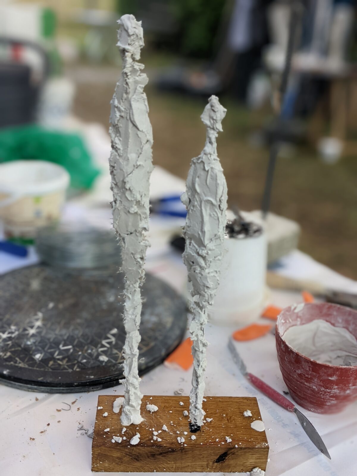 Zwei unfertige Skulpturen aus Ton auf einem Holzsockel, umgeben von verschiedenen Töpferwerkzeugen und Materialien.