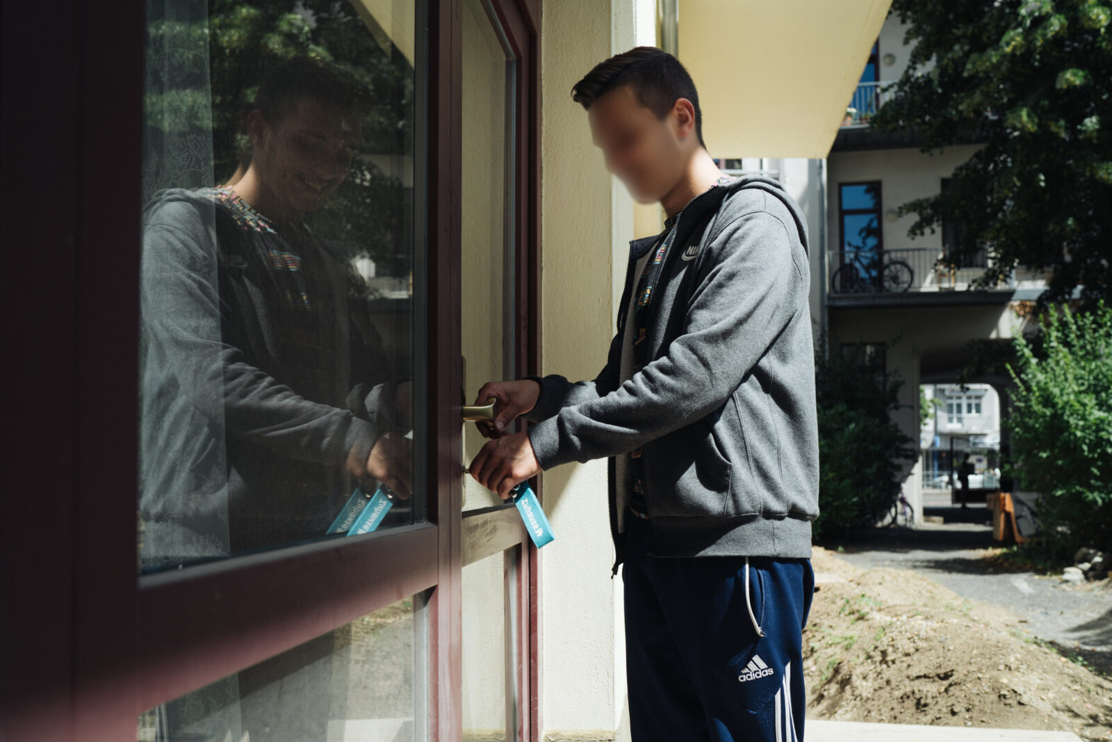 Ein Mann im Sportanzug öffnet eine Tür mit einem Schlüsselanhänger. Im Hintergrund sind Wohngebäude und grüne Bäume zu sehen.