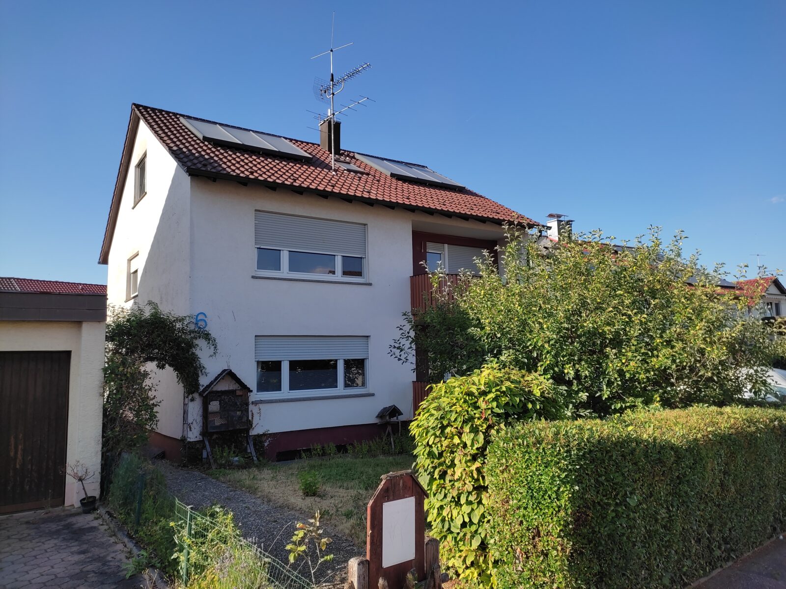 Zweistöckiges Wohnhaus mit rotem Dach und weißen Wänden, umgeben von einem gepflegten Garten mit grünen Büschen und Bäumen, an einem sonnigen Tag.