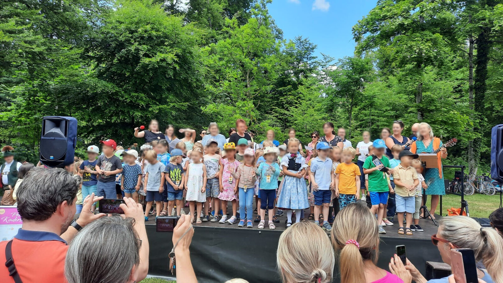 Eine Gruppe von Kindern steht auf einer Bühne im Freien, umgeben von Erwachsenen und Bäumen. Einige der Erwachsenen fotografieren die Szene. Ein Lautsprecher ist links auf der Bühne zu sehen, und eine Frau spielt Gitarre.