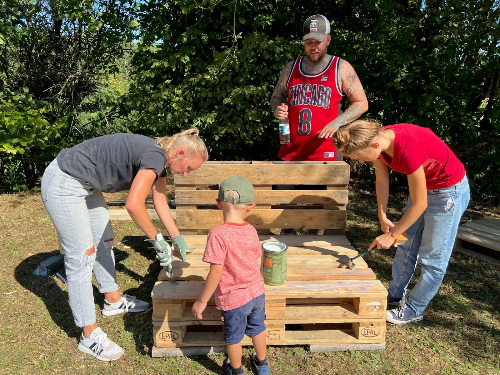 Eine Gruppe von Menschen, darunter ein Kind, streicht Holzpaletten im Freien bei sonnigem Wetter.