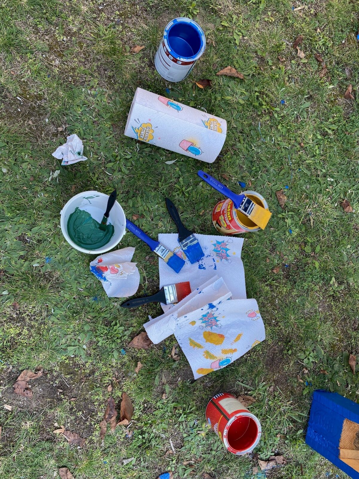 Mehrere Farbdosen und Pinsel liegen auf einer Grasfläche, umgeben von bemalten Papiertüchern.