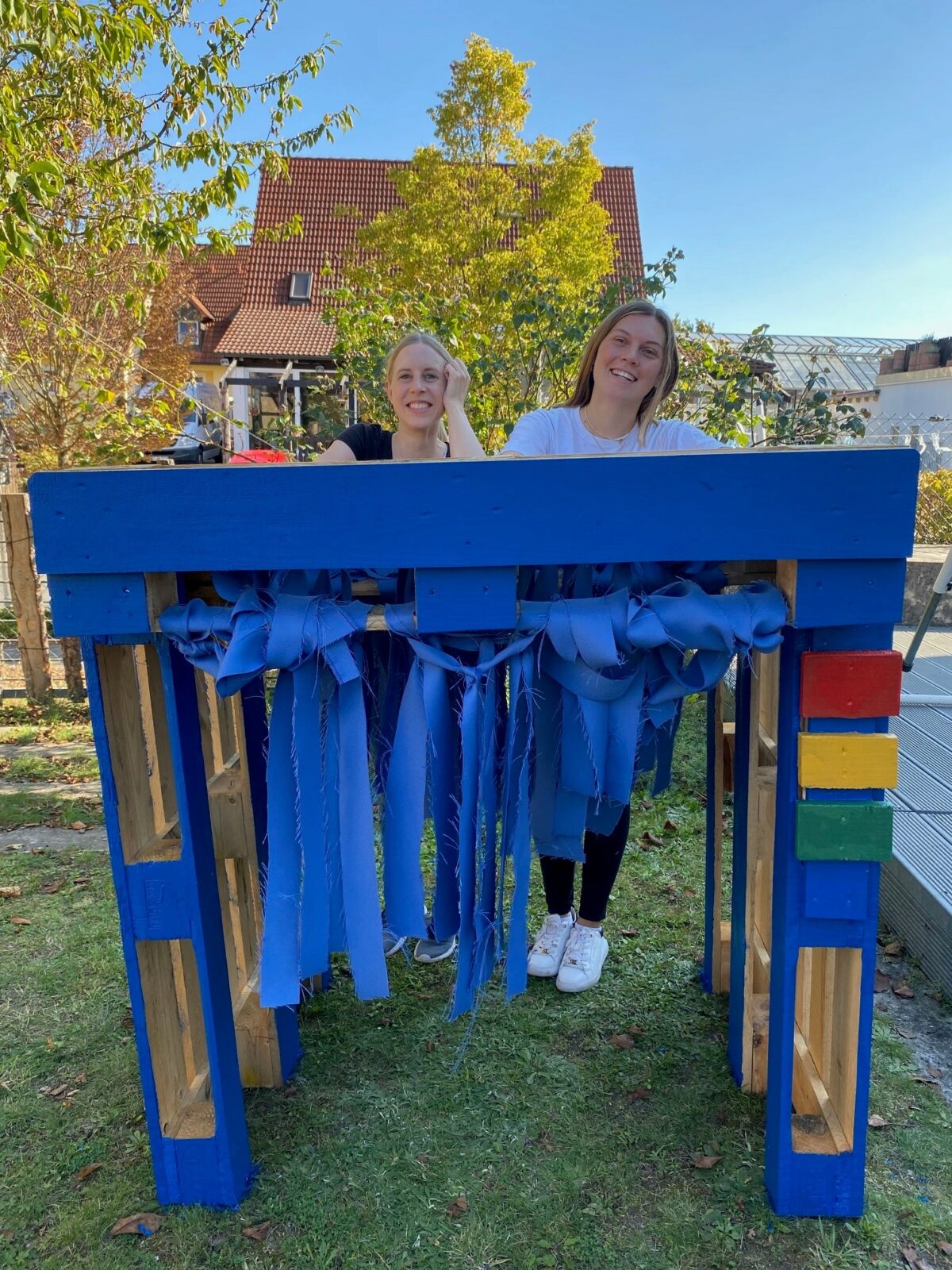 Zwei Frauen stehen hinter einem blauen Holztisch mit bunten Seitenstreifen und blauen Stoffbahnen, im Hintergrund sind Bäume und ein Haus zu sehen.