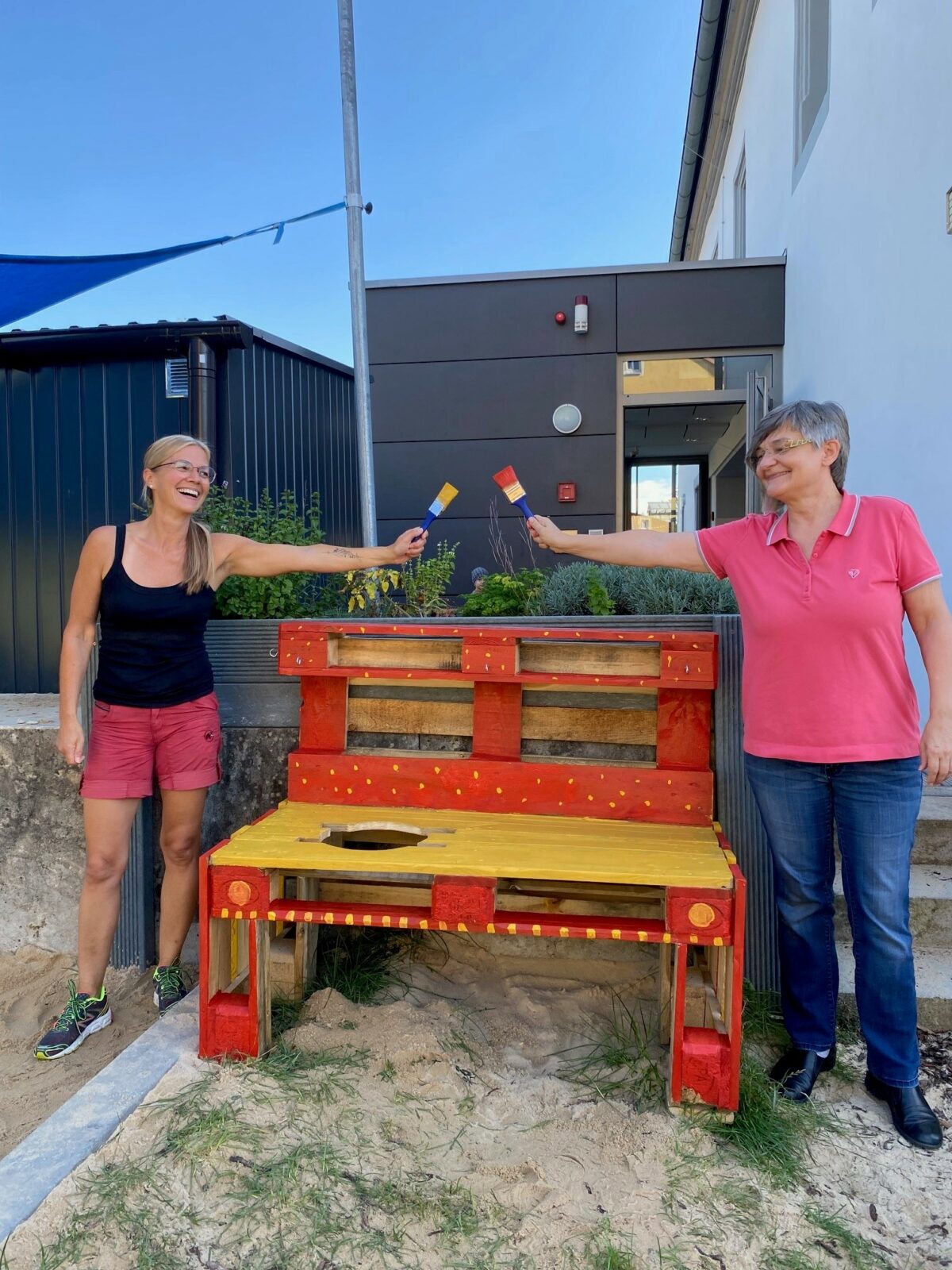 Zwei Frauen stehen neben einer bunt bemalten Holzbank im Freien und halten jeweils einen Pinsel hoch. Die Bank ist rot und gelb gestrichen und steht auf einem sandigen Boden vor einem modernen Gebäude.