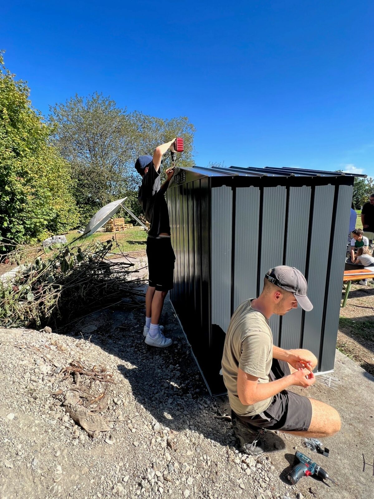 Zwei Personen arbeiten an der Montage eines kleinen, grauen Metallhäuschens im Freien. Eine Person steht aufrecht und befestigt ein Teil am Dach, während die andere Person kniet und Werkzeuge verwendet. Im Hintergrund sind Bäume und ein sonniger blauer Himmel zu sehen.
