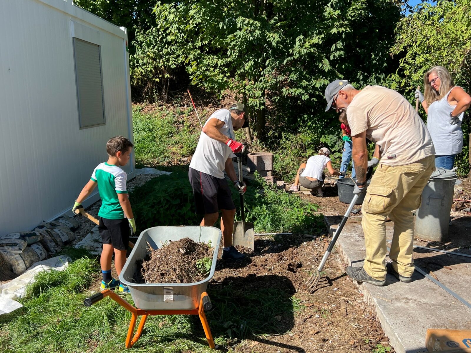 Eine Gruppe von Menschen arbeitet im Garten, ein Junge hält einen Spaten, während Erwachsene mit verschiedenen Gartenwerkzeugen den Boden bearbeiten. Im Vordergrund steht eine Schubkarre mit Kompost. Links ist ein weißes Gebäude zu sehen. Die Szene ist von grünen Bäumen umgeben.