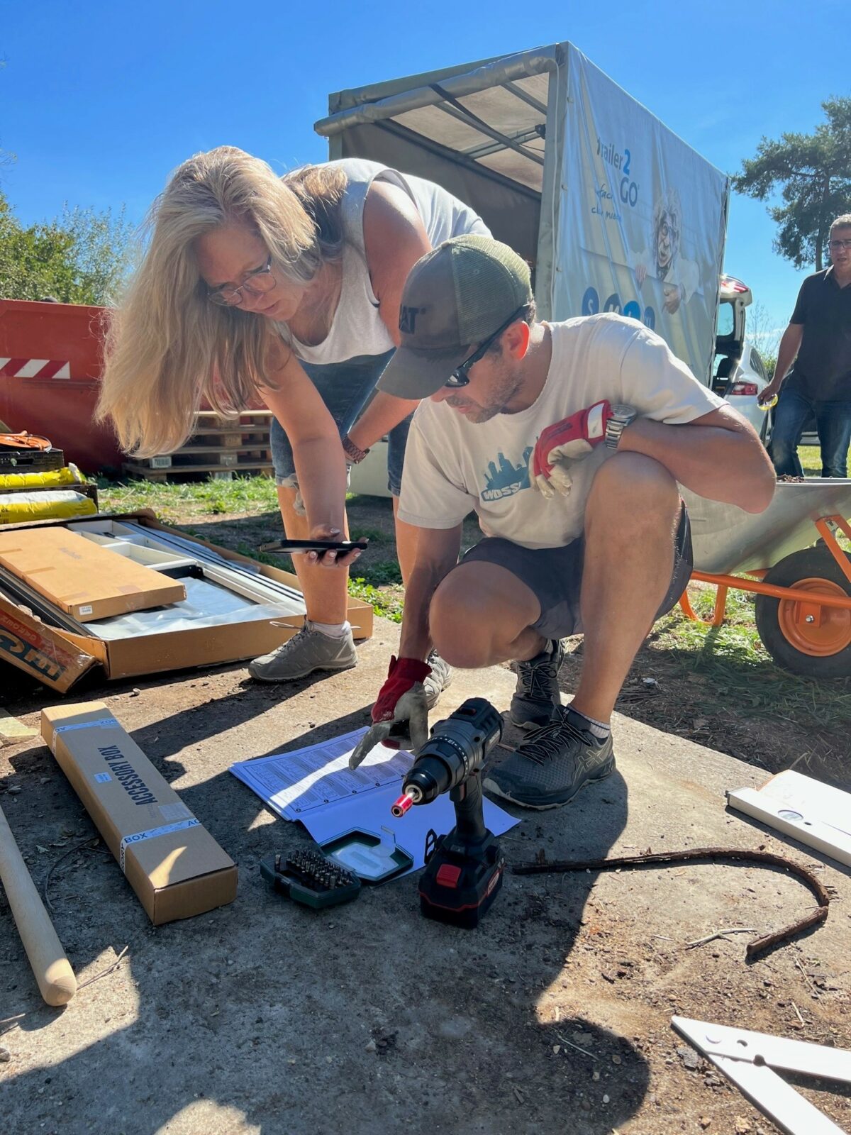 Zwei Personen arbeiten im Freien an einem Bauprojekt, umgeben von Werkzeugen und Materialien. Eine Person kniet und zeigt auf ein Dokument, während die andere Person daneben steht und auf das Handyschaut, welches sie in der Hand hält. Im Hintergrund sind ein Anhänger und eine Schubkarre zu sehen.