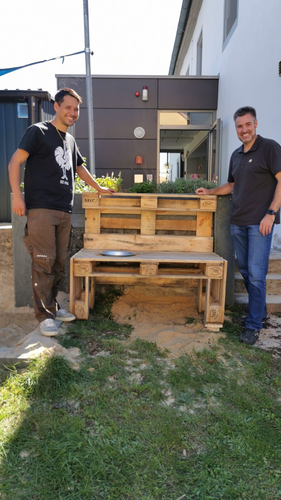 Zwei Personen stehen neben einer aus Holzpaletten gebauten Bank im Freien vor einem modernen Gebäude.