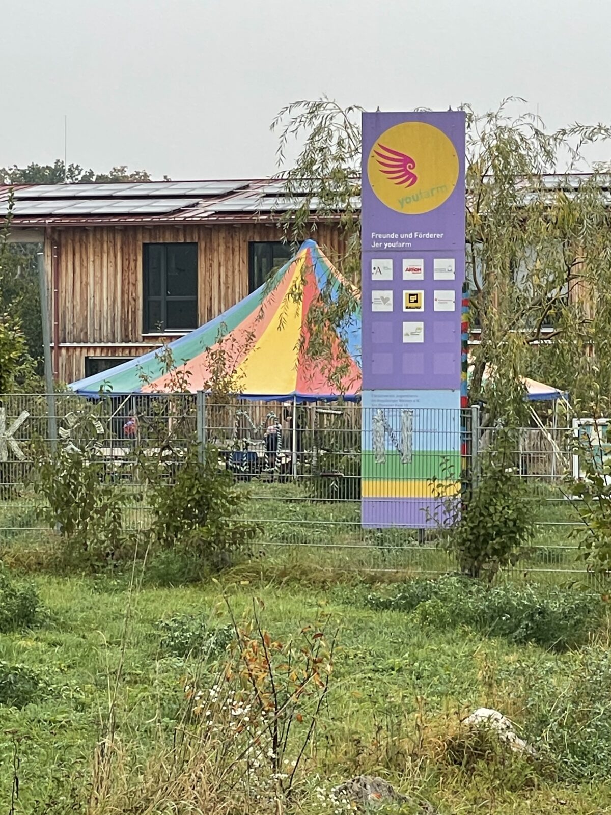 Ein großes Schild mit dem Logo und den Sponsoren von youfarm steht vor einem Gebäude aus Holz. Daneben ist ein buntes Zelt aufgebaut, umgeben von grünem Gras und Bäumen.