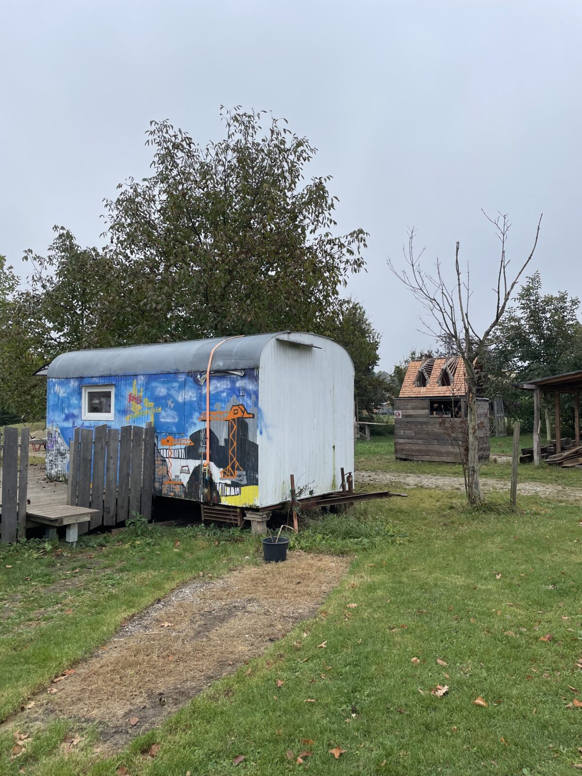Ein bunt bemalter Bauwagen steht auf einem grünen Grundstück, im Hintergrund sind Bäume und eine Holzhütte zu sehen.