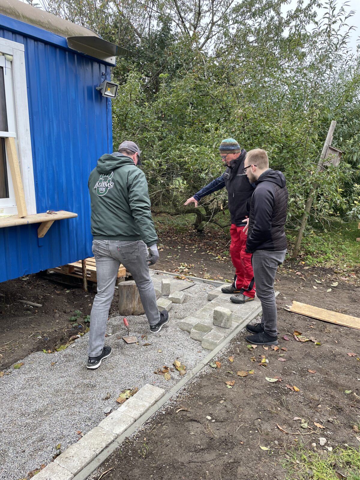 Drei Männer arbeiten gemeinsam an einem Gehweg aus Betonsteinen neben einem blauen Gebäude im Freien.