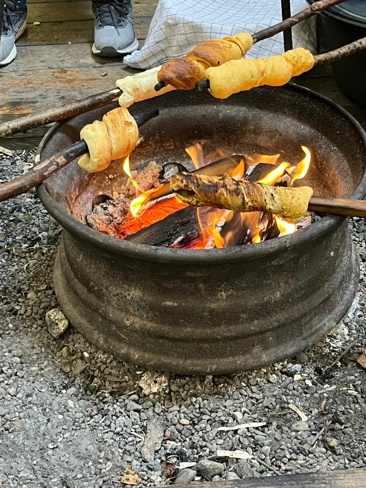 Stockbrot wird über einem offenen Feuer in einer Feuerschale gebacken, im Hintergrund sind Schuhe und ein Holzboden zu sehen.