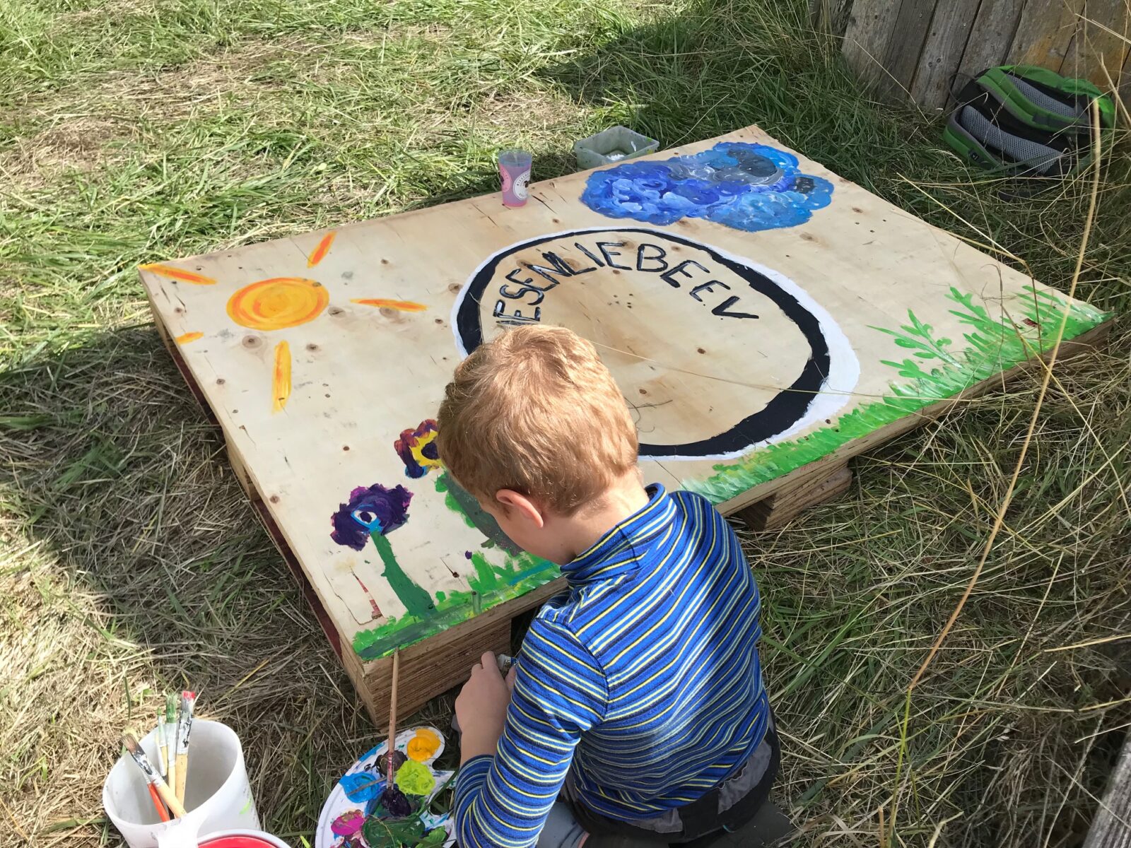 Ein Kind malt ein buntes Bild auf ein großes Holzbrett im Freien, umgeben von Gras. Das Bild zeigt eine Sonne, Blumen und den Schriftzug "Leben lieben". Neben dem Kind stehen Farbtöpfe und Pinsel.