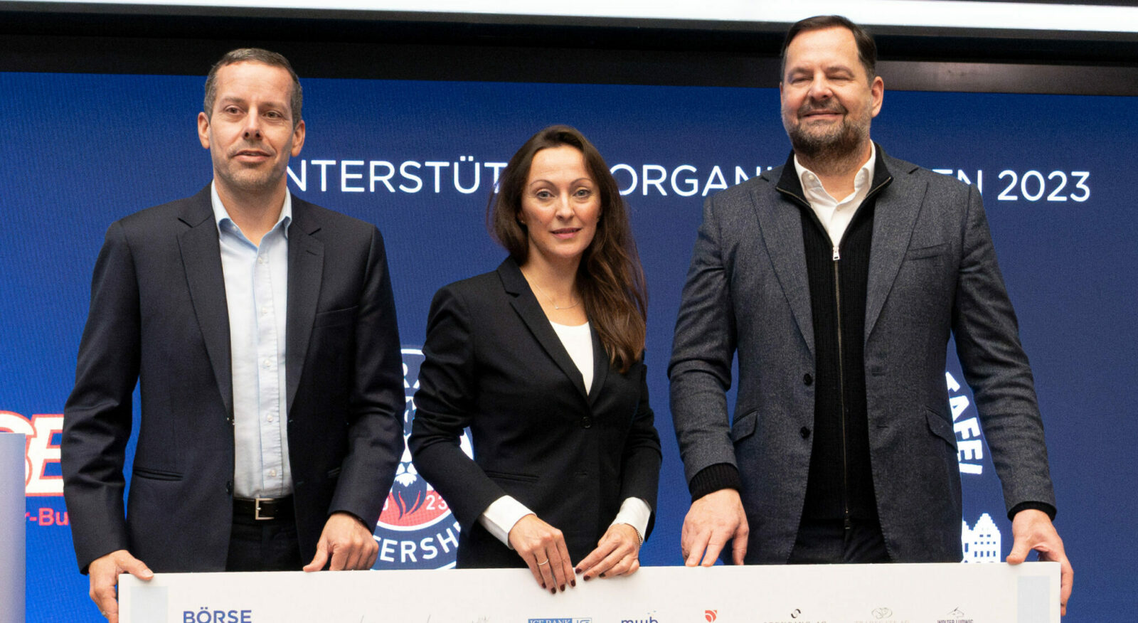 Drei Personen stehen nebeneinander vor einem blauen Hintergrund mit der Aufschrift "UNTERSTÜTZ ORGANISATIONEN 2023". Die Personen tragen Business-Kleidung und halten ein großes Schild.