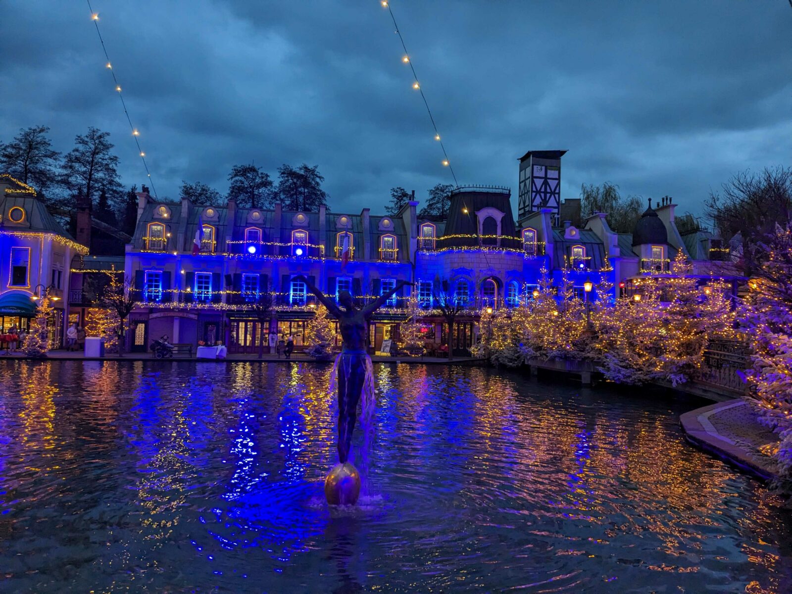 Beleuchtete Gebäude und Bäume am Ufer eines Gewässers in einem Freizeitpark bei Dämmerung, mit einer Statue im Vordergrund und Lichterketten, die über den Himmel gespannt sind.
