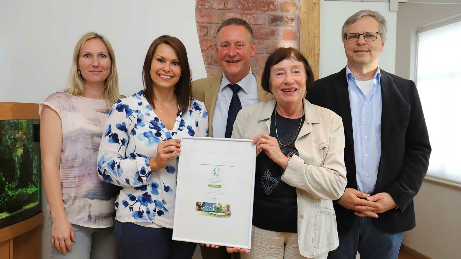 Eine Gruppe von Erwachsenen steht vor einer Wand und hält eine gerahmte Urkunde. Im Hintergrund ist ein Aquarium zu sehen.