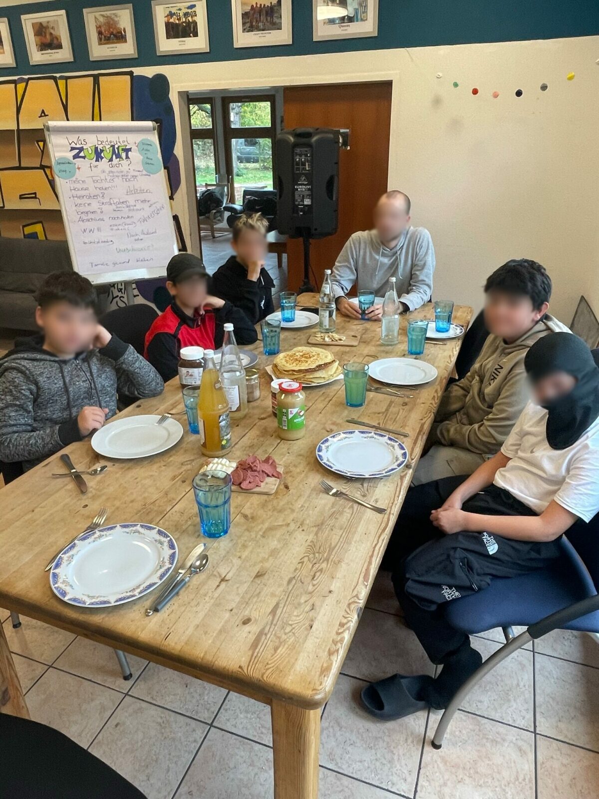 Eine Gruppe von jungen Menschen sitzt an einem Esstisch, der mit Tellern, Gläsern und Getränken gedeckt ist. Im Hintergrund steht ein Flipchart mit Notizen und eine Lautsprecherbox. An der Wand hängen mehrere gerahmte Bilder.