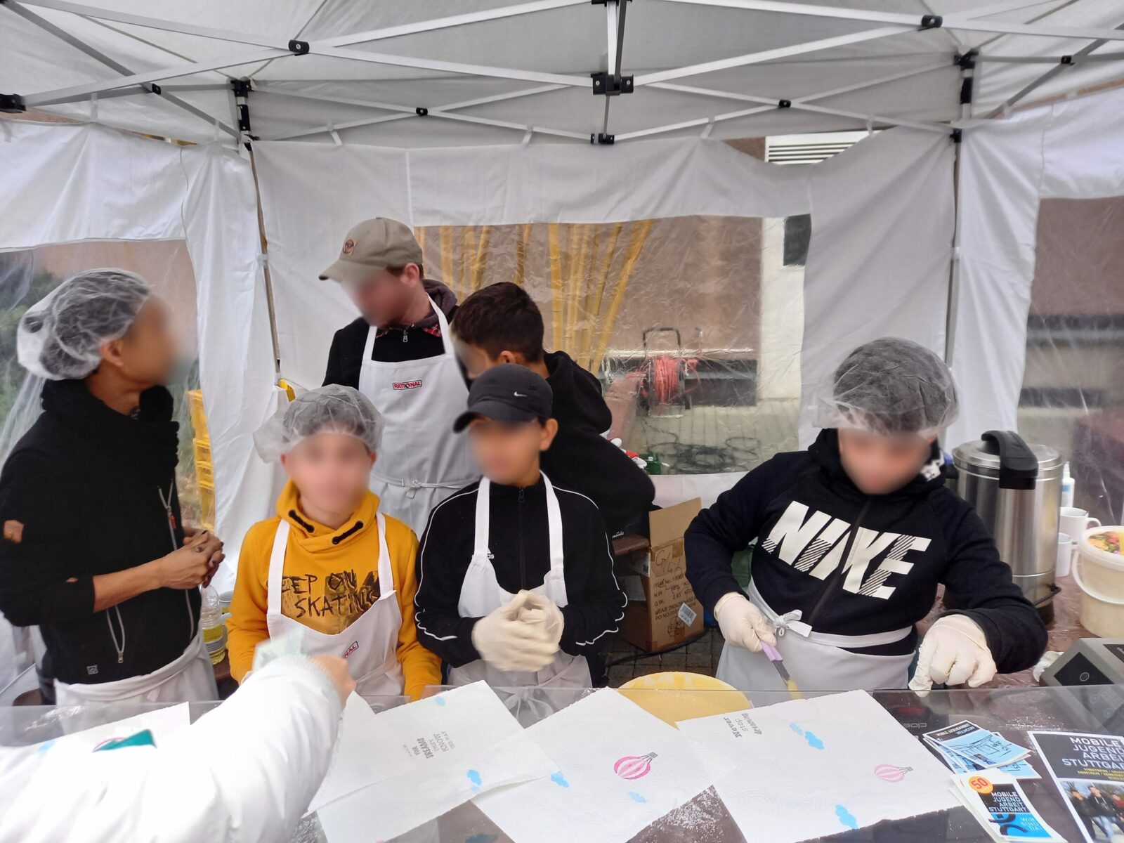 Gruppe von Jugendlichen in einem Zelt, die an einem Stand mit Essen arbeiten, während sie Schürzen und Haarnetze tragen.