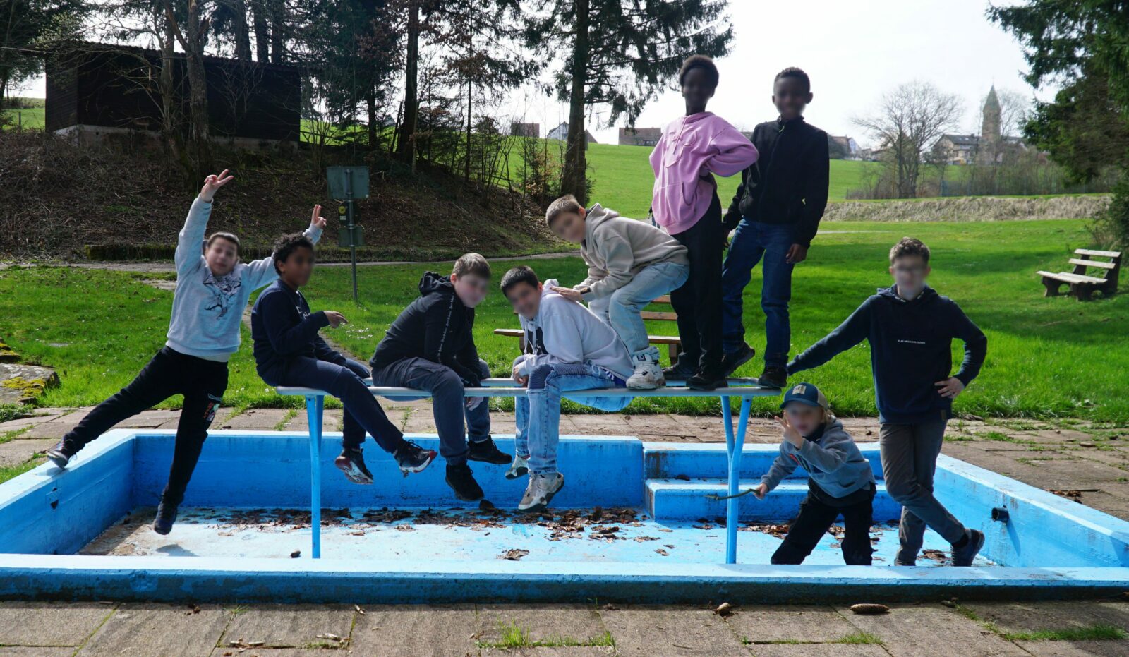 Eine Gruppe von Kindern spielt und posiert in einem leeren, blauen Wasserbecken in einem Park, umgeben von grüner Wiese und Bäumen.