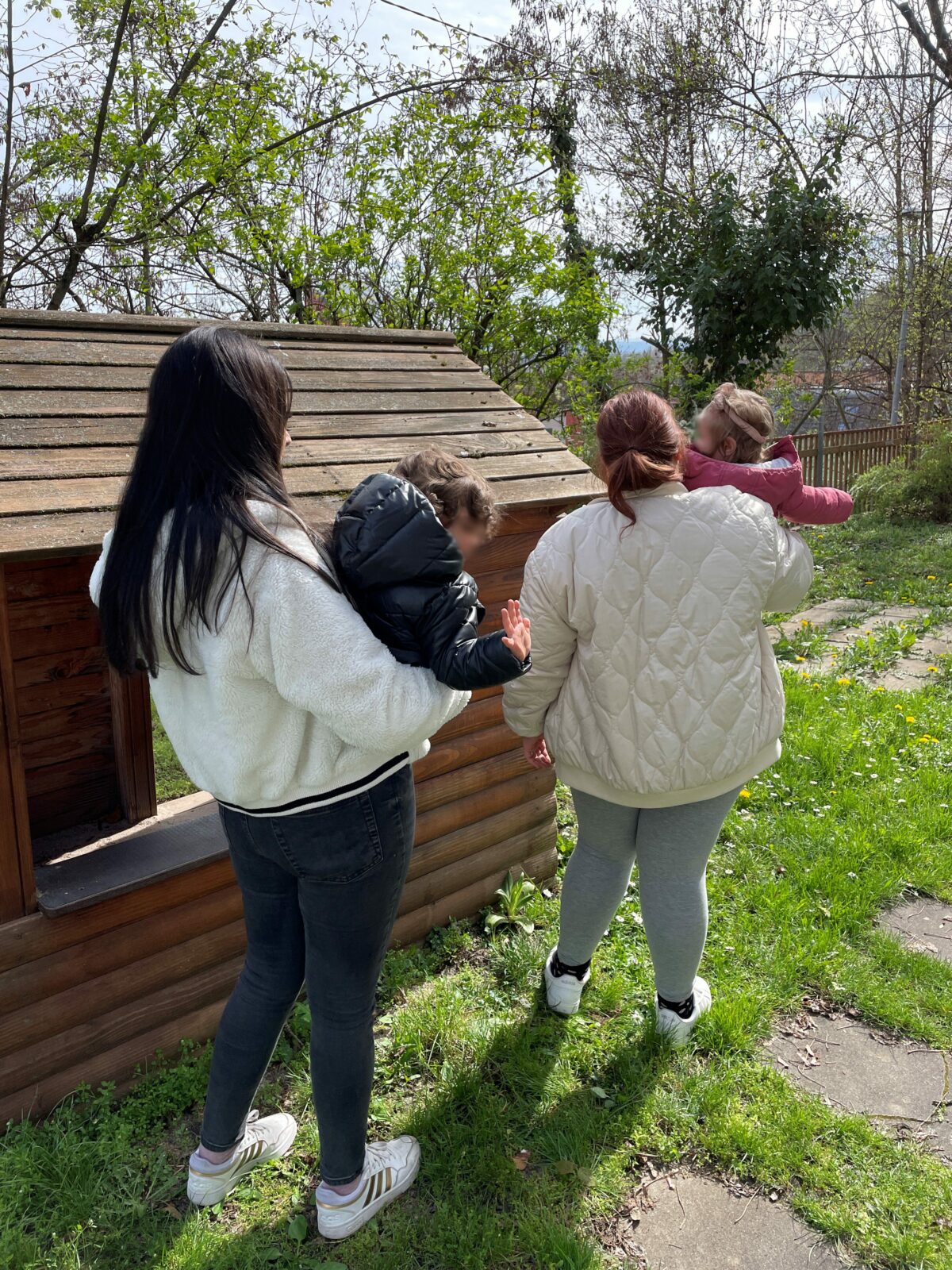 Das Bild zeigt zwei Frauen, die jeweils ein kleines Mädchen auf den Arm halten. Sie stehen in einem Garten vor einer kleinen Holzhütte.
