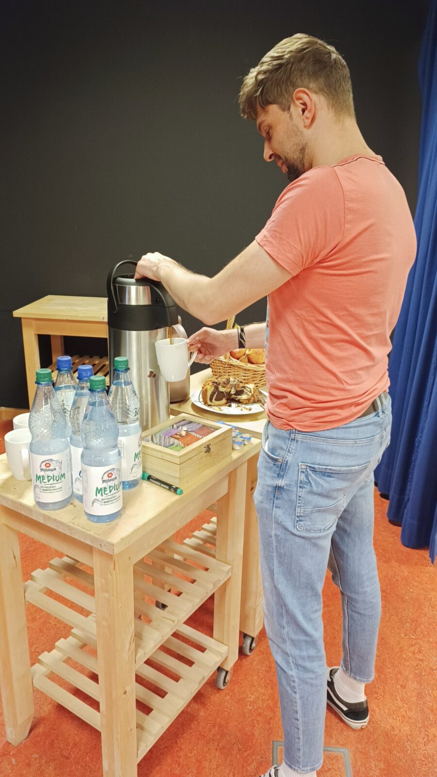 Auf einem Holztisch stehen einige Flaschenwasser eine Kaffekanne und Gebäck. Ein Mann steht davor und gießt sich aus der Kaffeekanne Kaffee in eine Tasse.