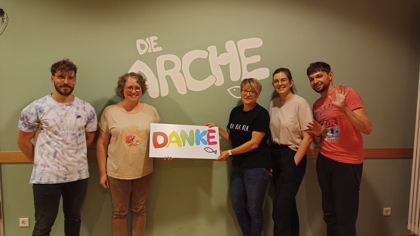 5 Erwachsene stehen vor einer Grünen Wand. Auf der grünen Wand steht "DIE ARCHE". Die zwei Erwachsenen in der Mitte halten ein Plakat mit der Aufschrift "DANKE". Sie schauen alle lächelnd und freundlich in die Kamera.