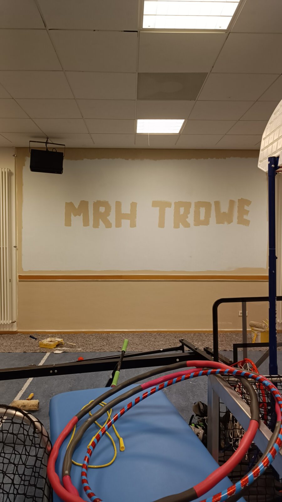 Das Bild zeigt eine frischgestrichene Sporthalle. Mit beiger Farbe wurde der Schriftzug MRH TROWE an die Wand gemalt.