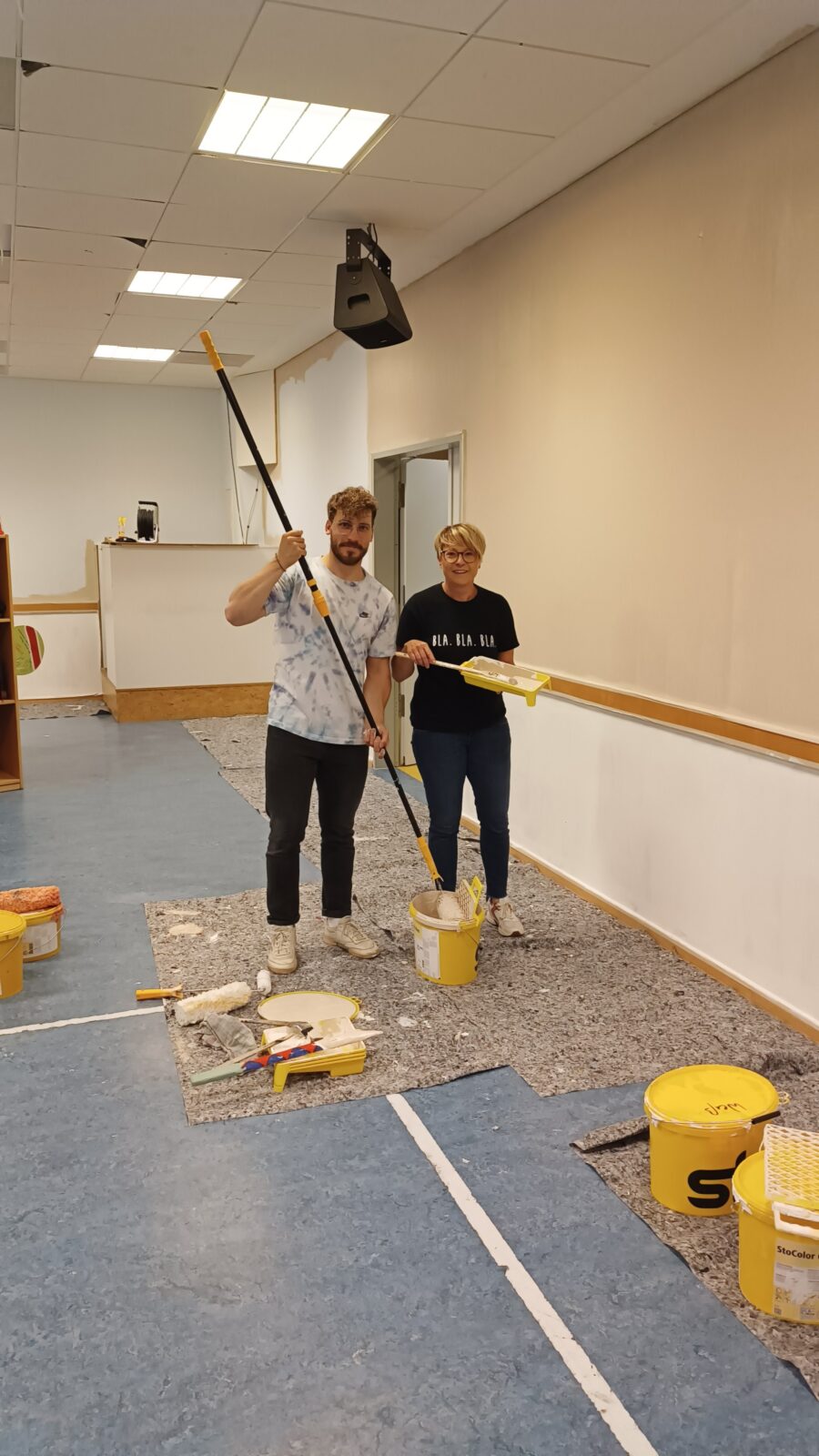 Eine Frau und ein Mann stehen nebeneinander in einem Raum. Der Mann hält eine Streichrolle in der Hand und tunkt diese in einen Eimer mit weißer Farbe. Die Frau hält eine Palette mit weißer Farbe in der einen Hand und in der anderen einen Pinsel.