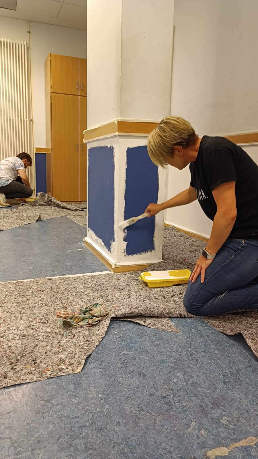 Eine Frau kniet auf dem Boden und sie hält einen Pinsel in der Hand. Sie bemalt mit dem Pinsel eine Säule in einem Raum mit weißer Farbe.