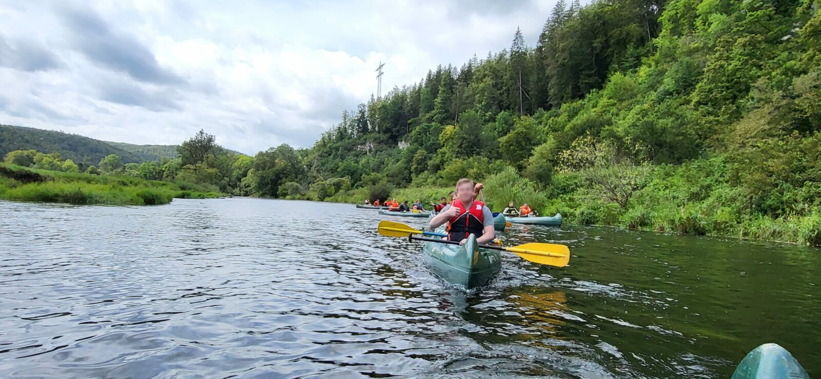 Zwei Menschen sitzen in einem grünen Kanu und paddeln durch einen Fluss, umgeben von bewaldeten Hügeln.