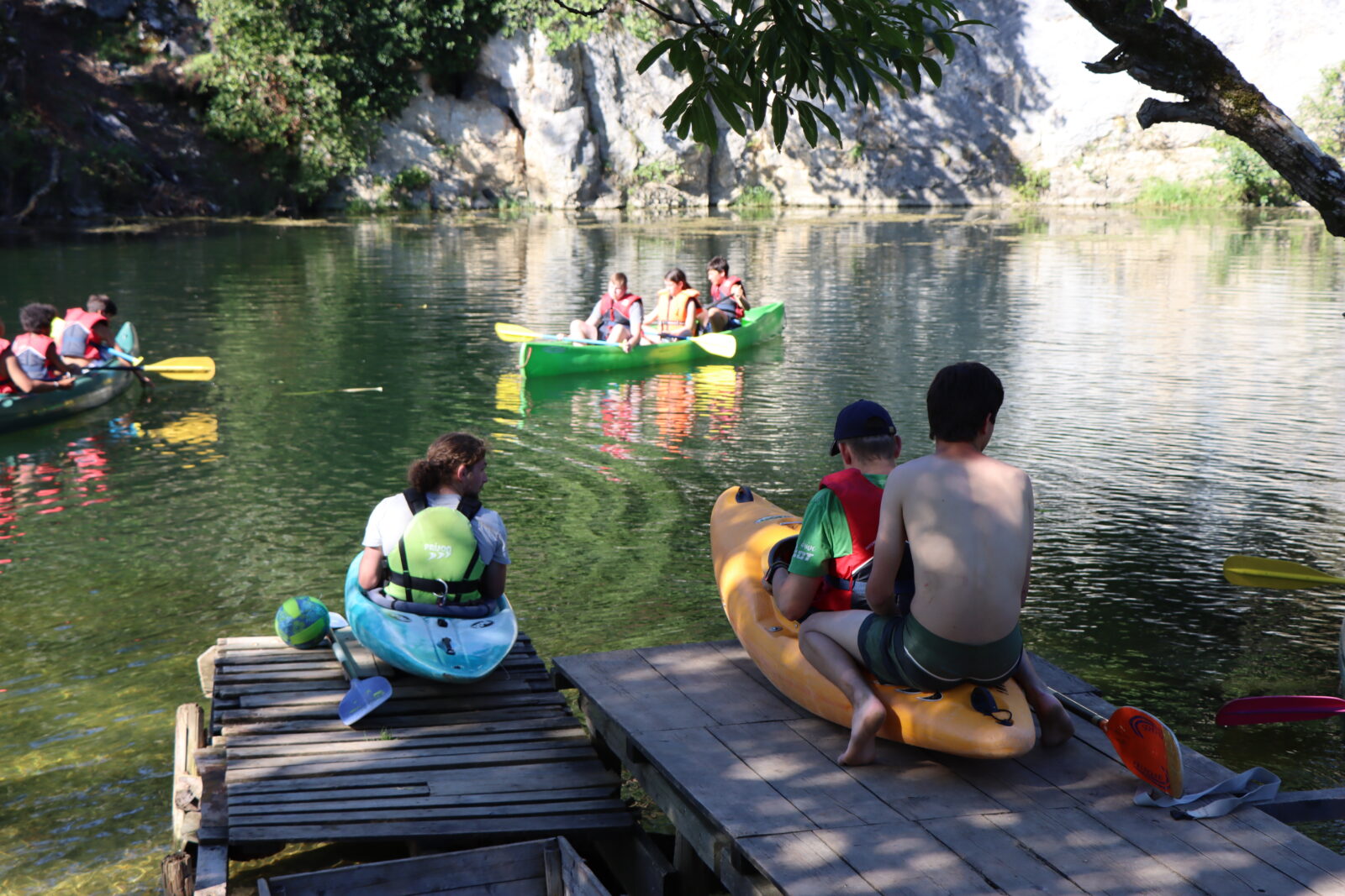 Mehrere Kinder sitzen verteilt in bunten Kanus und fahren über einen See. Drumherum sind Bäume und Berge zu sehen.