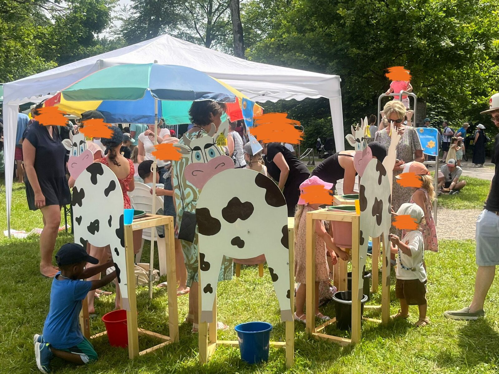 Kinder spielen unter einem weißen Pavillon mit Holzfiguren, die Kühen nachempfunden sind. Sie tragen bunte Kleidung und es handelt sich hierbei um ein Sommerfest.